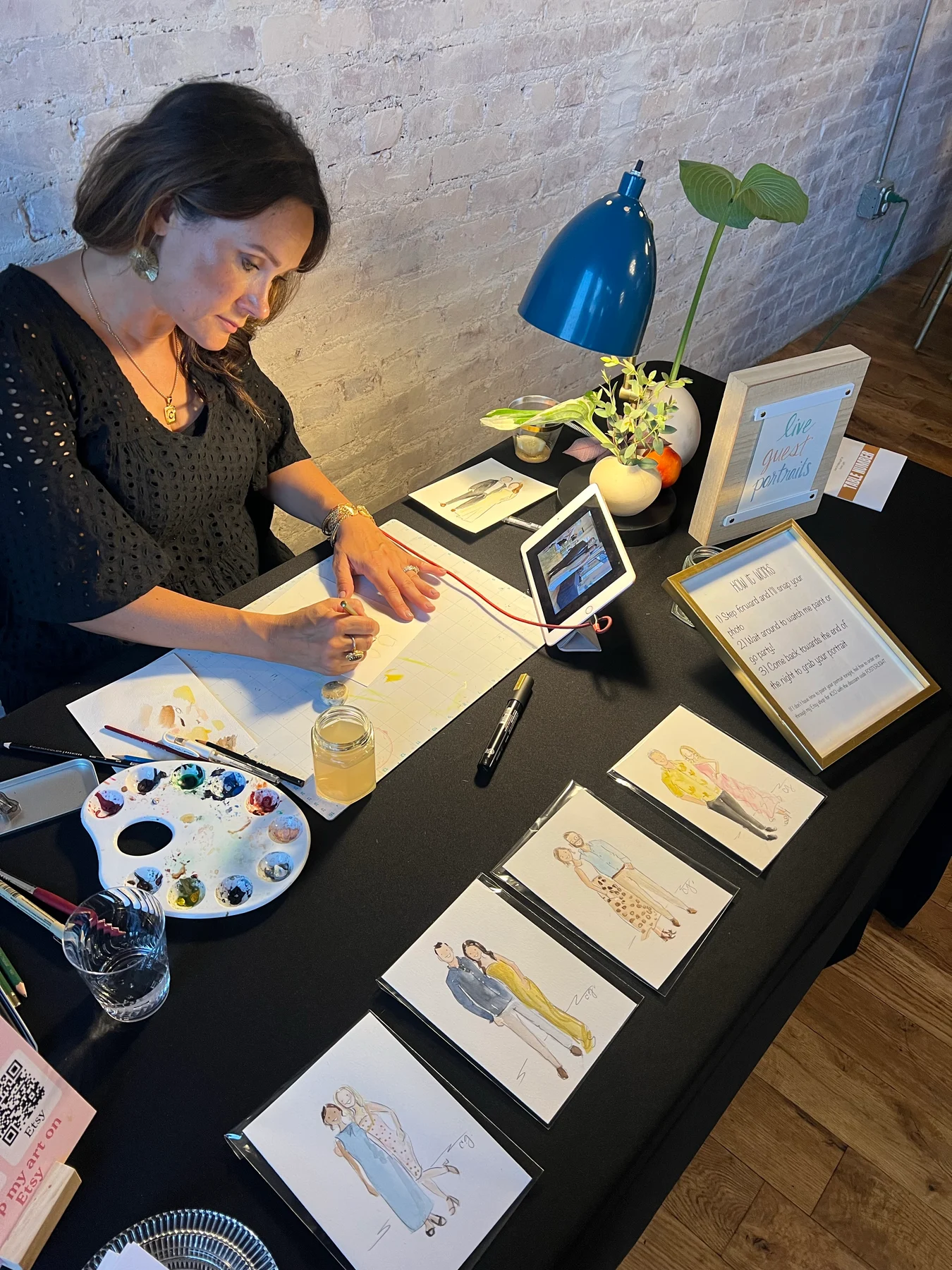 Carmen drawing portraits at an event, with her palette and finished paintings arranged on the table